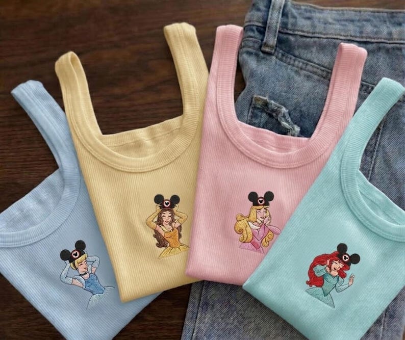 Puede incluir: Cuatro camisetas sin mangas acanaladas en colores pastel: azul claro, amarillo, rosa y turquesa. Cada camiseta presenta un bordado de una princesa de Disney con una diadema de orejas de Mickey Mouse. Las camisetas se muestran sobre un fondo de mezclilla.