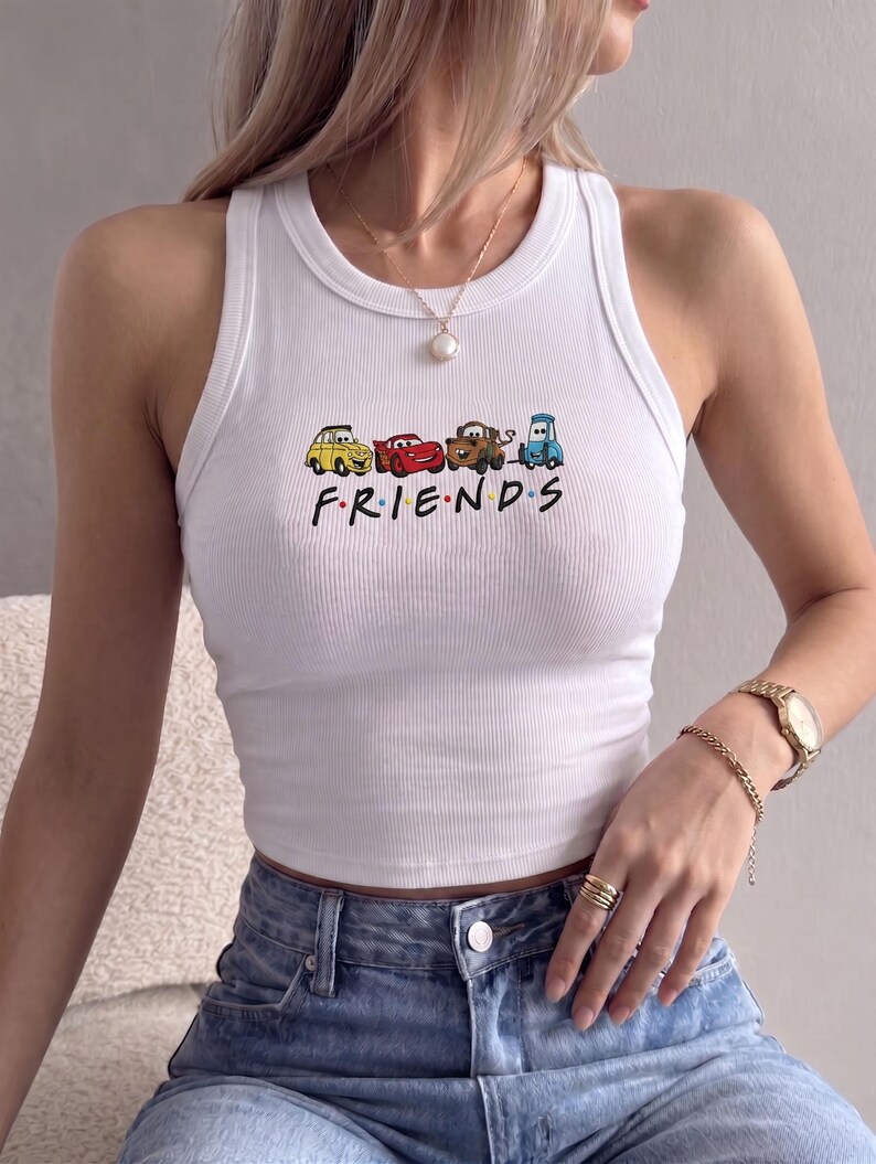 Op de afbeelding: Witte geribbelde tanktop met een kleurrijke afbeelding van cartoonauto's en het woord "FRIENDS" in het zwart. De top wordt gecombineerd met een blauwe jeans. Een gouden horloge en sieraden zijn ook zichtbaar.
