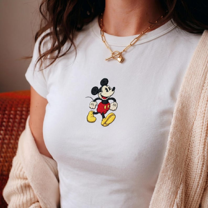 Puede incluir: Camiseta blanca con cuello redondo y dise&ntilde;o bordado de Mickey Mouse. El personaje de dibujos animados est&aacute; representado en negro, rojo, amarillo y blanco. La camiseta es de estilo cl&aacute;sico.