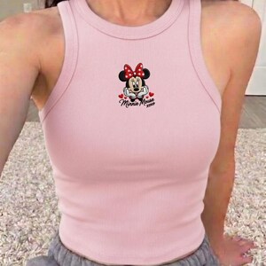 May include: A light pink ribbed tank top featuring a Minnie Mouse graphic. The graphic includes Minnie's face, a red bow, and the text "Minnie Mouse xoxo". The tank top has a round neckline and a racerback design.