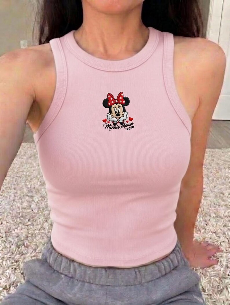 May include: A light pink ribbed tank top featuring a Minnie Mouse graphic. The graphic includes Minnie's face, a red bow, and the text "Minnie Mouse xoxo". The tank top has a round neckline and a racerback design.