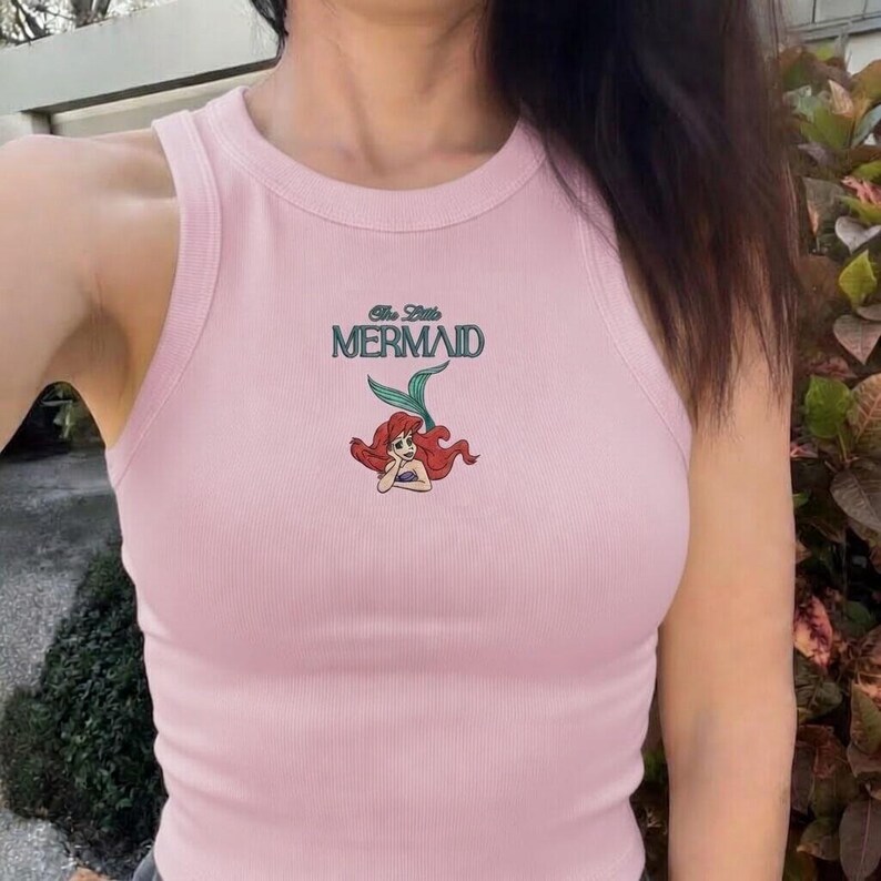 May include: A light pink ribbed tank top featuring the text "The Little Mermaid" above a cartoon illustration of Ariel. The top has a scoop neck and a sleeveless design, perfect for warm weather.