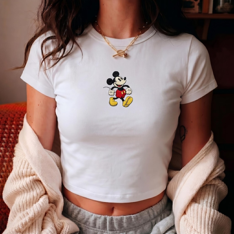 Puede incluir: Top corto blanco con una peque&ntilde;a imagen bordada de Mickey Mouse. El personaje de dibujos animados lleva pantalones cortos rojos, zapatos amarillos y guantes negros. La camiseta tiene mangas cortas y cuello redondo.