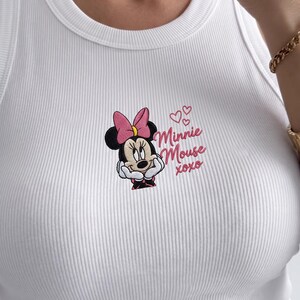 May include: White ribbed tank top featuring an embroidered Minnie Mouse design. The design includes Minnie's face, a pink bow, and the text "Minnie Mouse xoxo" in pink. The tank top has a scoop neckline.