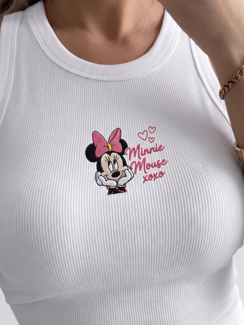 May include: White ribbed tank top featuring an embroidered Minnie Mouse design. The design includes Minnie's face, a pink bow, and the text "Minnie Mouse xoxo" in pink. The tank top has a scoop neckline.