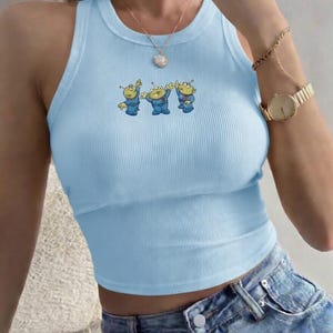 May include: Light blue ribbed tank top featuring a cartoon alien graphic. The sleeveless top has a round neckline and a cropped fit. The alien graphic is in shades of blue, yellow, and green. The top is paired with denim shorts.