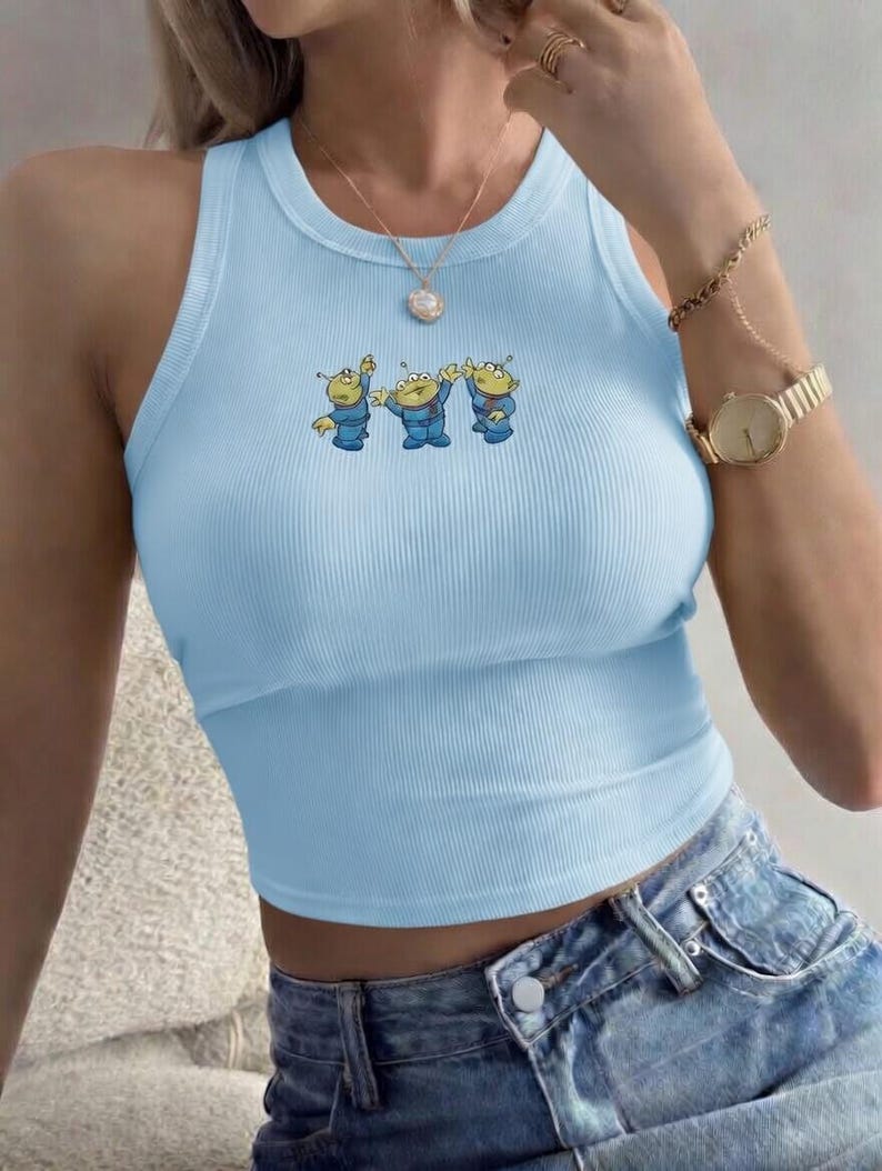 May include: Light blue ribbed tank top featuring a cartoon alien graphic. The sleeveless top has a round neckline and a cropped fit. The alien graphic is in shades of blue, yellow, and green. The top is paired with denim shorts.