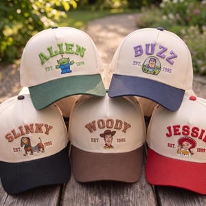 May include: Five baseball caps with cream-colored crowns and colored brims. Each cap features embroidered character names, character illustrations, and the text EST. 1995 or 1999. The caps feature Alien, Buzz, Slinky, Woody, and Jessie designs.