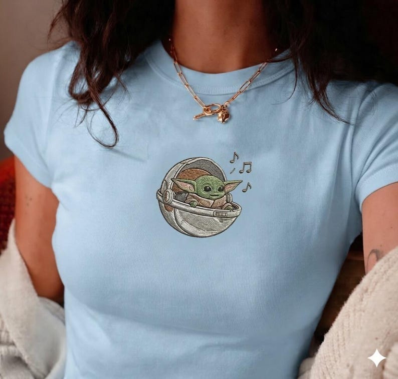K&ouml;nnte beinhalten: Hellblaues T-Shirt mit einem gestickten Bild eines gr&uuml;nen Alien-Charakters in einer Kapsel, begleitet von Noten. Das Design befindet sich auf der Brust. Das T-Shirt hat kurze &Auml;rmel und einen Rundhalsausschnitt.