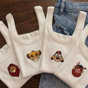 May include: Four white ribbed tank tops with embroidered Lion King character faces: Simba, Timon, Pumbaa, and Mufasa. The tank tops are displayed on a pair of blue jeans.