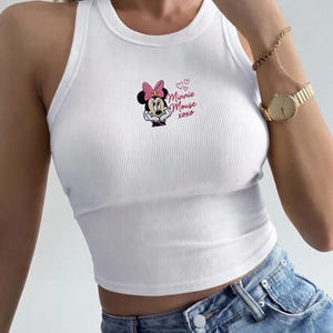 May include: White ribbed tank top featuring a Minnie Mouse graphic with a pink bow and the text "Minnie Mouse xoxo". The top is a cropped style and is paired with blue denim jeans. A gold watch and bracelet are also visible.