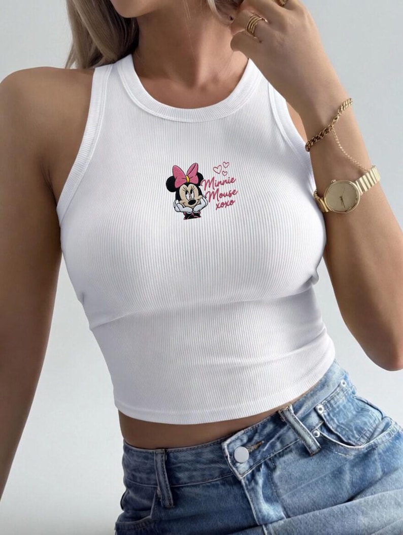 May include: White ribbed tank top featuring a Minnie Mouse graphic with a pink bow and the text "Minnie Mouse xoxo". The top is a cropped style and is paired with blue denim jeans. A gold watch and bracelet are also visible.