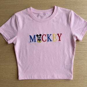 以下が含まれることがあります： 丸首と半袖の明るいピンク色のクロップドトップ。 「ミッキー」の文字は、前面に青、黒、赤、黄色の糸で刺繍されており、「i」の代わりに漫画のキャラクターが使用されています。
