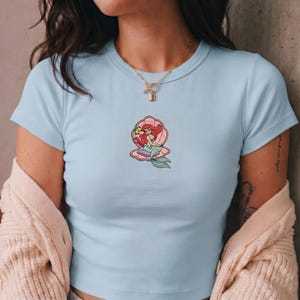 May include: Light blue t-shirt featuring an embroidered design of a mermaid sitting in a seashell. The mermaid has red hair and a green tail. The seashell is pink and purple.