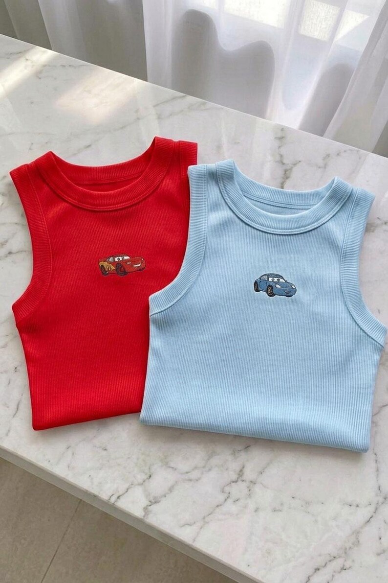 May include: Two ribbed tank tops, one red and one light blue, each with a small embroidered car design. The tops are sleeveless and have a round neckline. The red top has a yellow and red car, and the blue top has a blue car.