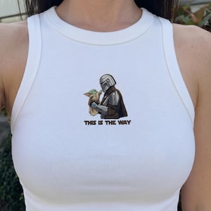 Peut inclure: D&eacute;bardeur blanc c&ocirc;tel&eacute; avec un motif du personnage de Mandalorien tenant Grogu, avec le texte "THIS IS THE WAY" en dessous. Le design utilise des tons de marron, vert, argent et noir.