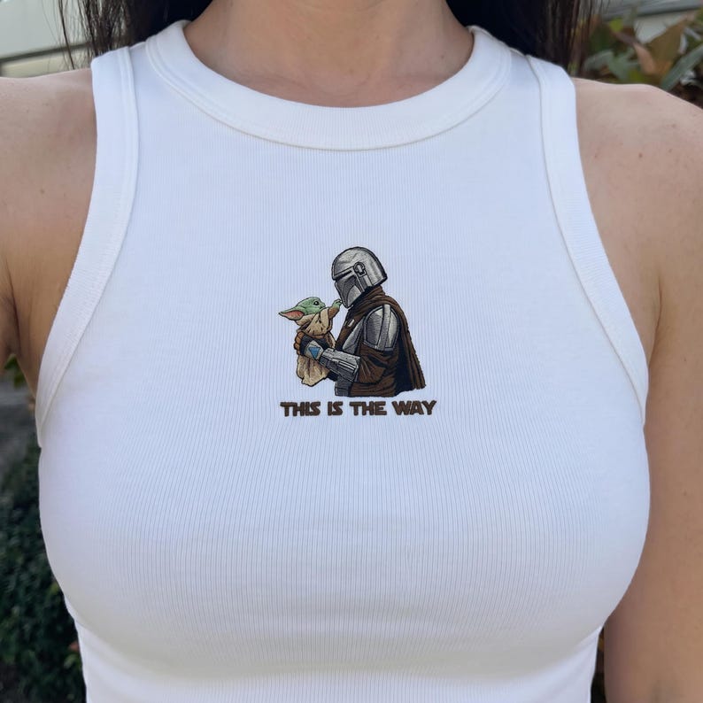Peut inclure: D&eacute;bardeur blanc c&ocirc;tel&eacute; avec un motif du personnage de Mandalorien tenant Grogu, avec le texte "THIS IS THE WAY" en dessous. Le design utilise des tons de marron, vert, argent et noir.
