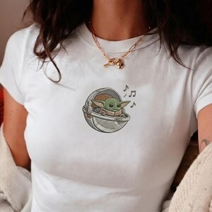K&ouml;nnte beinhalten: Wei&szlig;es T-Shirt mit einem gestickten Bild eines gr&uuml;nen Alien-Charakters in einer silbernen Kapsel, mit Noten. Der Charakter hat gro&szlig;e Ohren und ein kleines L&auml;cheln. Das T-Shirt hat kurze &Auml;rmel.