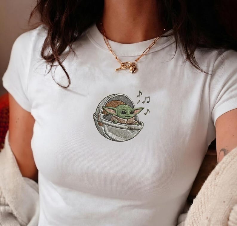 K&ouml;nnte beinhalten: Wei&szlig;es T-Shirt mit einem gestickten Bild eines gr&uuml;nen Alien-Charakters in einer silbernen Kapsel, mit Noten. Der Charakter hat gro&szlig;e Ohren und ein kleines L&auml;cheln. Das T-Shirt hat kurze &Auml;rmel.