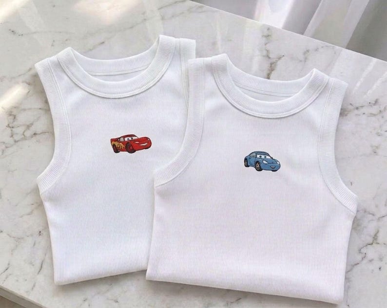 May include: Two white ribbed tank tops with embroidered cartoon car designs. One tank top features a red car, the other a blue car. Both tops have a round neckline and sleeveless design, perfect for warm weather.