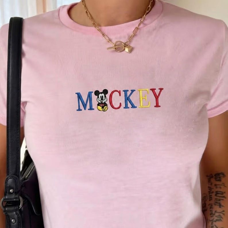 以下が含まれることがあります： 水色Tシャツに、青、赤、黄色の刺繍で「MCKEY」の文字が施され、中央には漫画のネズミのキャラクターが描かれています。Tシャツは丸いネックラインと半袖です。