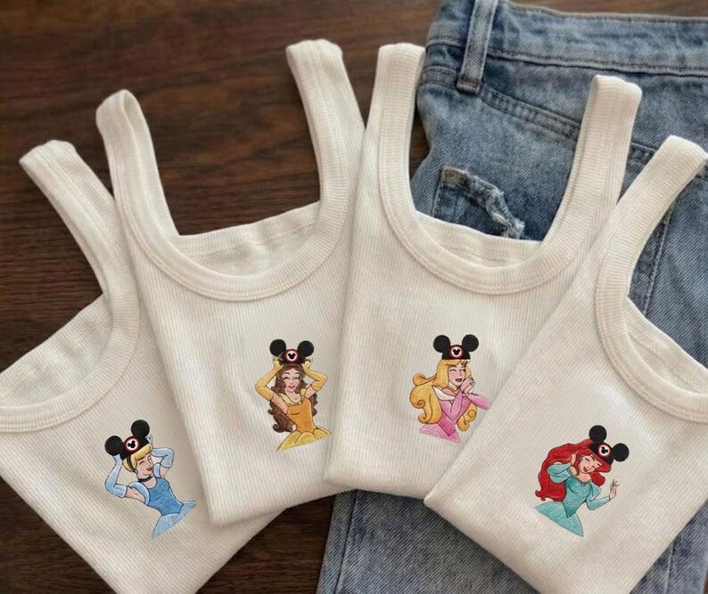 Puede incluir: Cuatro camisetas sin mangas blancas acanaladas con bordados de princesas de Disney. Cada camiseta sin mangas presenta una princesa diferente con una diadema de orejas de Mickey Mouse. Las camisetas sin mangas se muestran sobre un fondo de mezclilla.