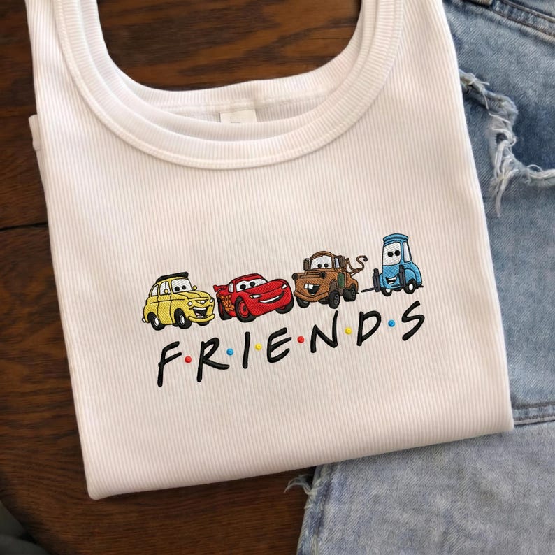 Op de afbeelding: Witte geribbelde tanktop met een geborduurd ontwerp van tekenfilmauto's en het woord "FRIENDS" in zwarte letters. De auto's zijn geel, rood, bruin en blauw. Het woord "FRIENDS" heeft gekleurde stippen.
