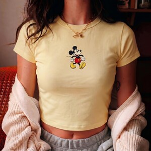 Puede incluir: Un top corto amarillo claro con un dise&ntilde;o de Mickey Mouse. La camiseta de manga corta tiene cuello redondo. El dise&ntilde;o de Mickey Mouse es en negro, rojo, amarillo y blanco. La parte superior es de material suave.