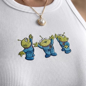 May include: White ribbed tank top featuring embroidered cartoon aliens in blue spacesuits. A gold chain necklace with a pearl pendant is also visible. The tank top is a casual, everyday wear item.