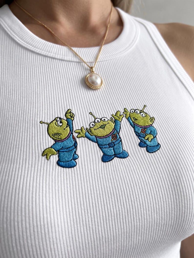 May include: White ribbed tank top featuring embroidered cartoon aliens in blue spacesuits. A gold chain necklace with a pearl pendant is also visible. The tank top is a casual, everyday wear item.