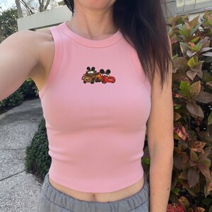 May include: Pink ribbed tank top featuring a cartoon graphic of two Cars characters, Mater and Lightning McQueen. The sleeveless top has a round neckline and is made of a soft, ribbed fabric. The graphic is in the center of the chest.