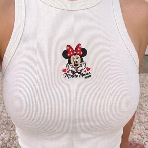May include: White ribbed tank top featuring an embroidered Minnie Mouse design. The design includes Minnie's face, a red bow, and the text "Minnie Mouse xoxo" with heart accents. The tank top has a round neckline and a sleeveless design.