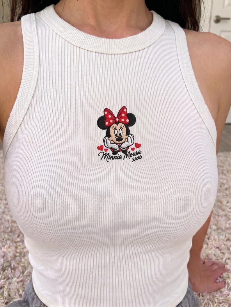 May include: White ribbed tank top featuring an embroidered Minnie Mouse design. The design includes Minnie's face, a red bow, and the text "Minnie Mouse xoxo" with heart accents. The tank top has a round neckline and a sleeveless design.