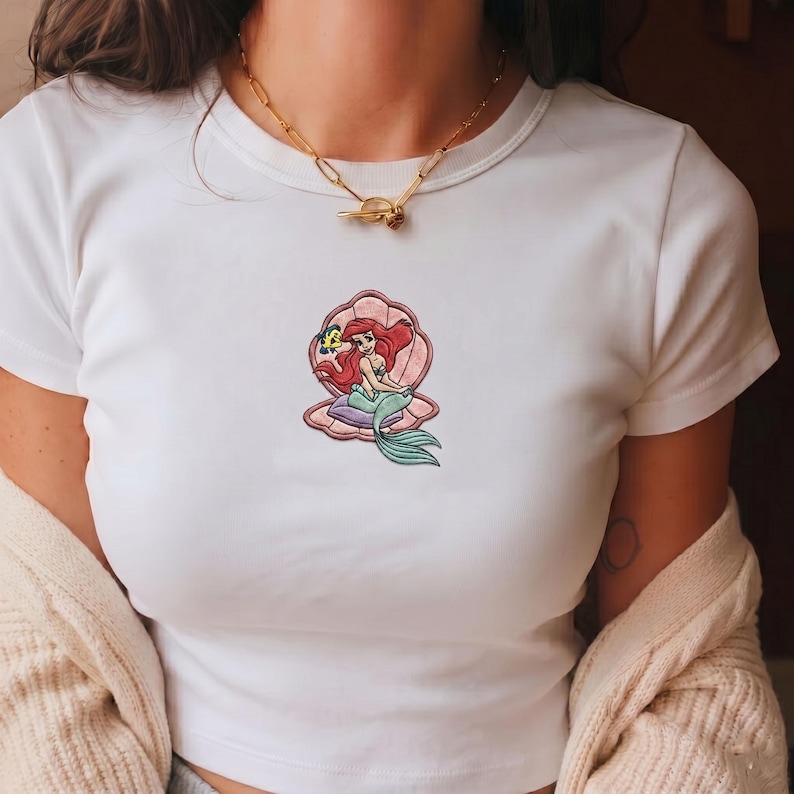 May include: White t-shirt featuring a colorful embroidered design of a mermaid sitting in a seashell. The mermaid has red hair and a green tail. The seashell is pink and the design includes a yellow fish.