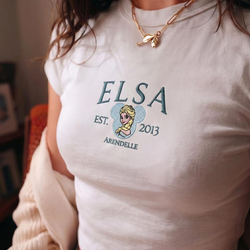 Puede incluir: Una camiseta blanca de manga corta con un dise&ntilde;o bordado que incluye el texto ELSA, EST. 2013 y ARENDELLE en hilo azul verdoso oscuro. El gr&aacute;fico central incluye una peque&ntilde;a ilustraci&oacute;n de un personaje rubio dentro de una forma de oreja.