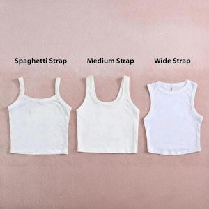 May include: Three white crop tops with different strap styles: spaghetti strap, medium strap, and wide strap. The tops are displayed on a pink background, showcasing the variations in design. The text labels each style.