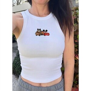 May include: White ribbed tank top featuring a cartoon graphic of two Cars characters, Mater and Lightning McQueen. The graphic is in shades of brown and red. The tank top is sleeveless.