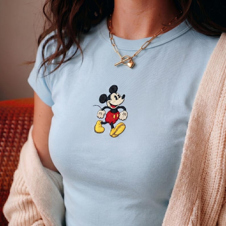 Puede incluir: Camiseta azul claro con un bordado de Mickey Mouse. El personaje de dibujos animados lleva pantalones cortos rojos, zapatos amarillos y guantes blancos. Tambi&eacute;n se ve un collar dorado con un colgante en forma de coraz&oacute;n.
