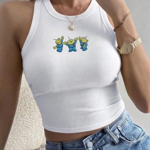 May include: White ribbed tank top featuring a cartoon graphic of three green aliens in blue suits. The sleeveless top has a crew neck and a cropped fit. The aliens are in a row, with their arms raised. The top is paired with blue denim jeans.