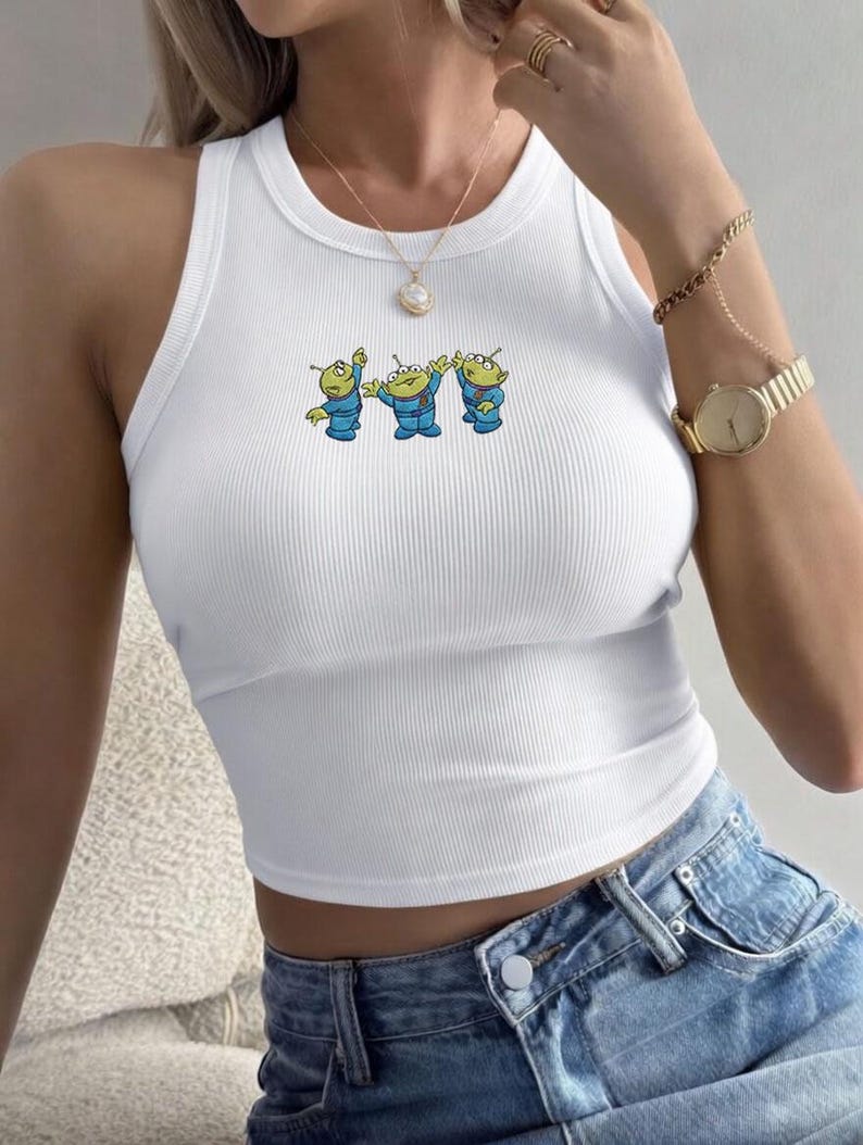 May include: White ribbed tank top featuring a cartoon graphic of three green aliens in blue suits. The sleeveless top has a crew neck and a cropped fit. The aliens are in a row, with their arms raised. The top is paired with blue denim jeans.