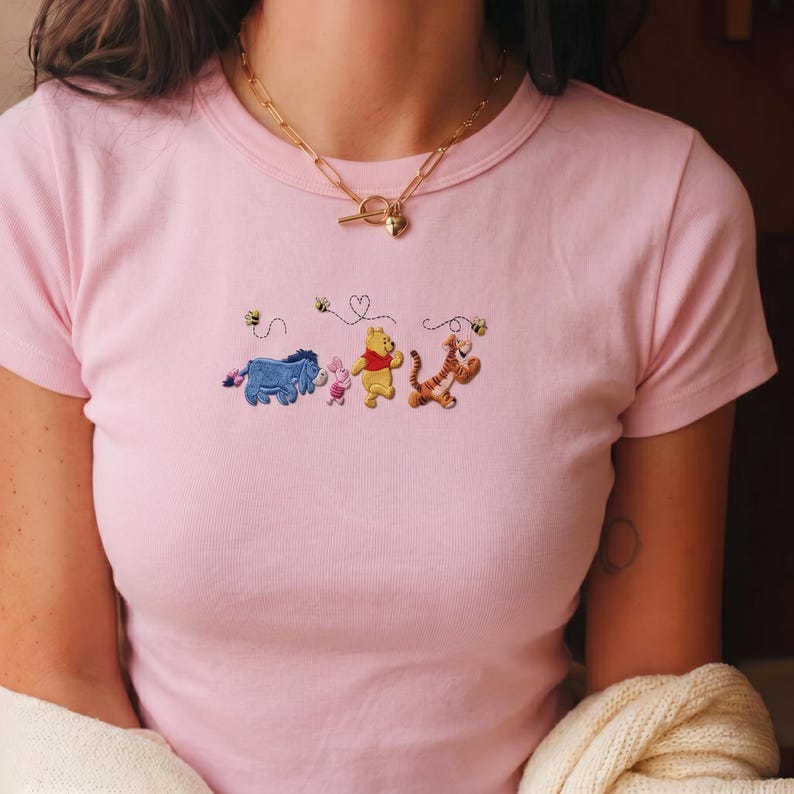 May include: A light pink t-shirt with embroidered characters from a children's story. The design features a blue donkey, a pink pig, a yellow bear, and an orange tiger, with bees and hearts. A gold necklace is worn.