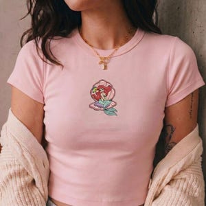 May include: A light pink t-shirt featuring a graphic of a mermaid inside a seashell. The mermaid has red hair and a teal tail. The t-shirt has a crew neck and short sleeves.