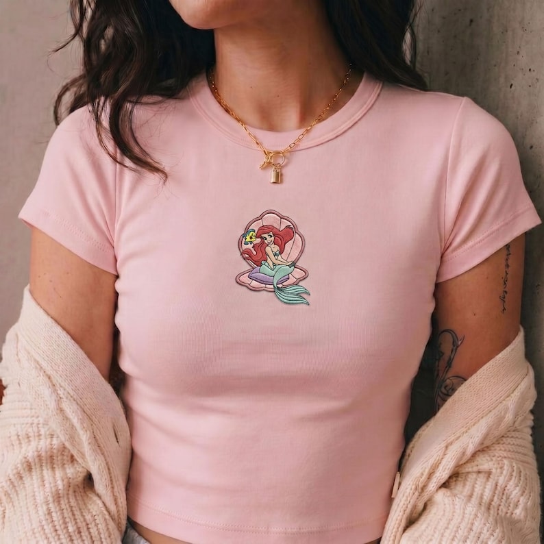 May include: A light pink t-shirt featuring a graphic of a mermaid inside a seashell. The mermaid has red hair and a teal tail. The t-shirt has a crew neck and short sleeves.