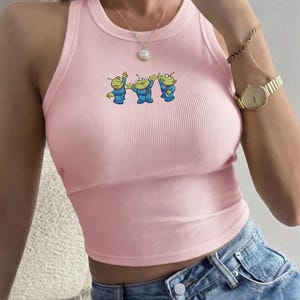 May include: A light pink ribbed crop top with a crew neck and a cartoon graphic of three green aliens in blue suits. The top is sleeveless and has a fitted silhouette. The aliens are in a playful pose.