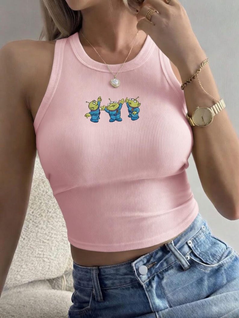 May include: A light pink ribbed crop top with a crew neck and a cartoon graphic of three green aliens in blue suits. The top is sleeveless and has a fitted silhouette. The aliens are in a playful pose.