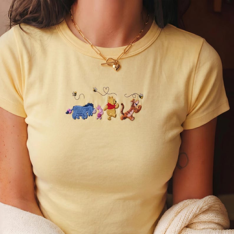 May include: A light yellow t-shirt with embroidered images of Winnie the Pooh characters: Eeyore, Piglet, Winnie the Pooh, and Tigger. The shirt has a crew neck and short sleeves. A gold-colored necklace with a heart pendant is worn.