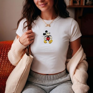 Puede incluir: Top corto blanco con un gr&aacute;fico de Mickey Mouse. El personaje de dibujos animados se representa con pantalones cortos rojos, zapatos amarillos y guantes negros. Una prenda casual para el d&iacute;a a d&iacute;a.
