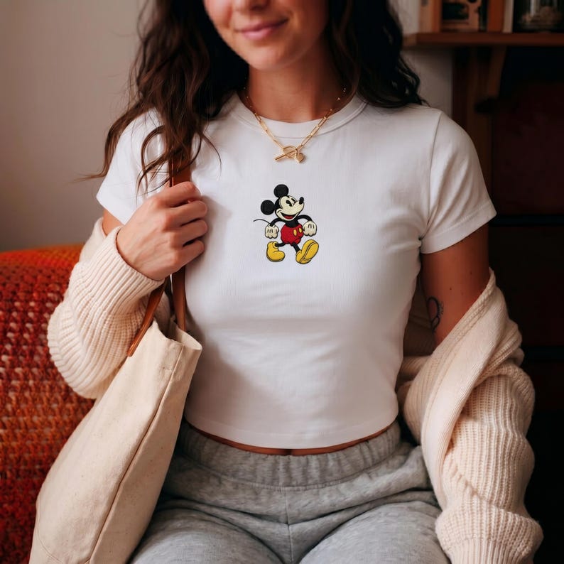 Puede incluir: Top corto blanco con un gr&aacute;fico de Mickey Mouse. El personaje de dibujos animados se representa con pantalones cortos rojos, zapatos amarillos y guantes negros. Una prenda casual para el d&iacute;a a d&iacute;a.