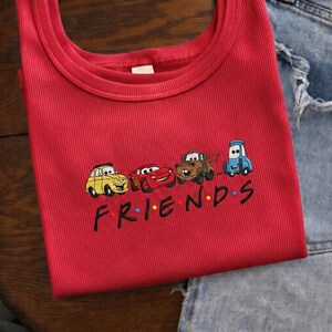 Op de afbeelding: Een rode geribbelde tanktop met een geborduurd ontwerp van tekenfilmauto's en het woord "FRIENDS" in het zwart. De auto's zijn geel, rood, bruin en blauw. De tanktop is opgevouwen.