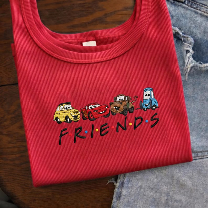 Op de afbeelding: Een rode geribbelde tanktop met een geborduurd ontwerp van tekenfilmauto's en het woord "FRIENDS" in het zwart. De auto's zijn geel, rood, bruin en blauw. De tanktop is opgevouwen.
